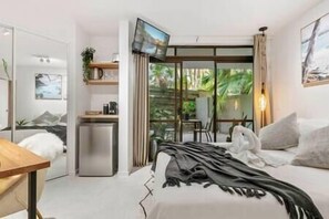 Un dormitorio moderno con cama, un escritorio pequeño, un refrigerador y una puerta corrediza de vidrio que da a un balcón.
