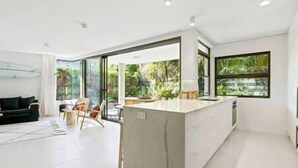 A modern kitchen with a large island, a dining area with chairs, and a view of greenery through large windows.