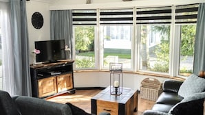 A living room with a TV on a wooden cabinet, a black sofa, a coffee table, and large windows with a view of greenery. - Chalet in Veluwe Near Kootwijkerzand (Putten)