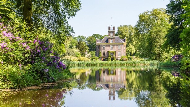 A historic building with a symmetrical facade, a central tower, and a reflection in a calm water body surrounded by lush greenery and flowering plants. - Chalet in Veluwe Near Kootwijkerzand (Putten)