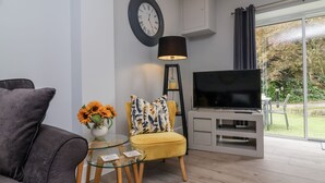 A modern living room with a sofa, a yellow chair, a glass coffee table, a TV on a white entertainment unit, and a large window with a view of greenery. - Beach Hideaway (Bideford)