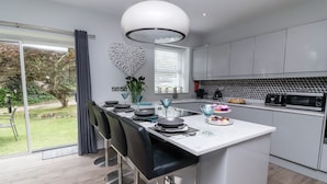 A modern kitchen with a central island, bar stools, a dining table, and a view of the garden. - Beach Hideaway (Bideford)