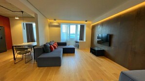 A modern living room with a sectional sofa, a dining table with chairs, a flat-screen TV mounted on the wall, and a large window providing natural light.