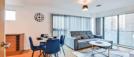Ein modernes Wohnzimmer mit Essbereich, einem grauen Sofa, einem runden Couchtisch und einem großen Fenster mit Blick auf die Stadt.