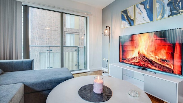 A modern living room with a large TV displaying a fireplace, a grey sofa, and a glass table with a candle. - City Retreat in Manchester (Manchester)