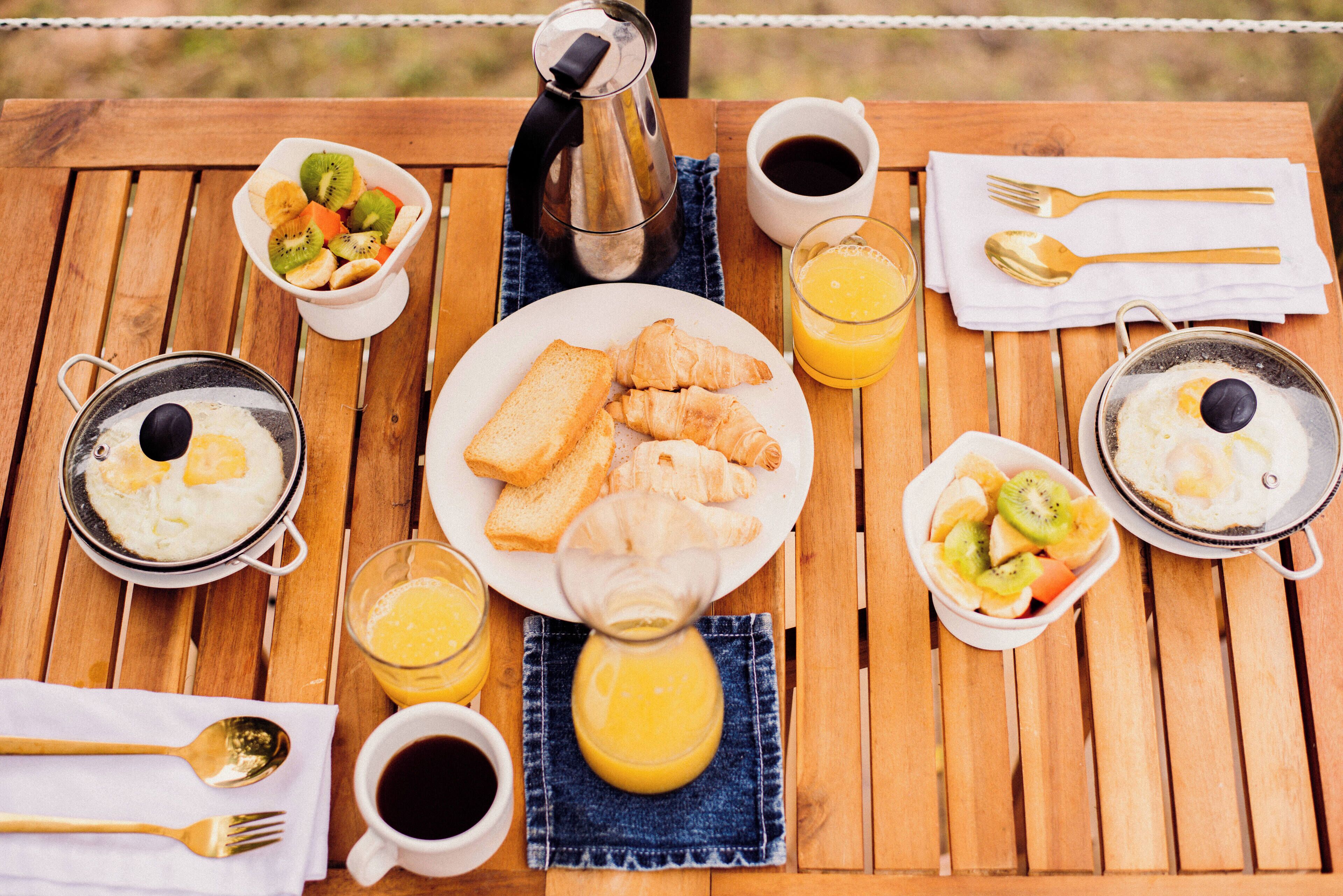 Una mesa de madera con alimentos para el desayuno, como cruasanes, tostadas, huevos y fruta. También hay tazas de café y zumo de naranja.