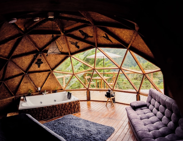 Interior de una cúpula geodésica con piso de madera, una bañera grande, un sofá y vistas a un paisaje verde exuberante a través de un amplio ventanal.