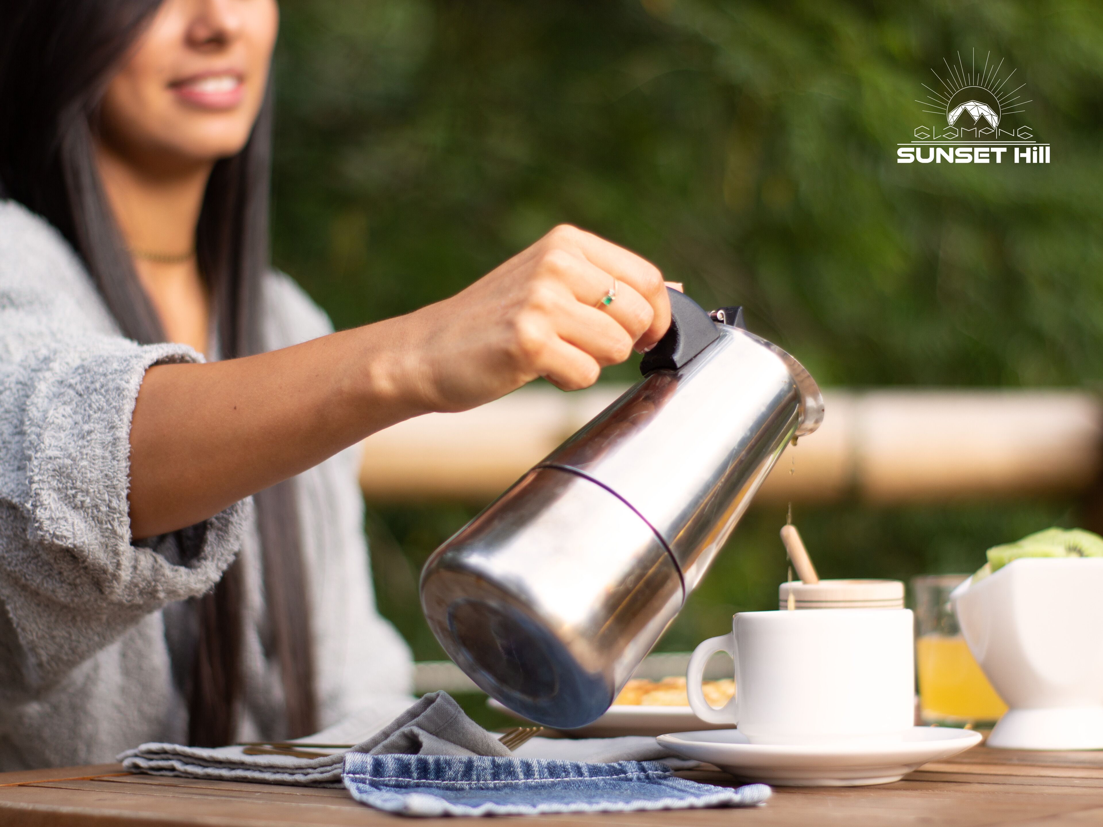 A person pouring a beverage from a stainless steel kettle into a white cup, with a plate of food and a glass of juice on the table.