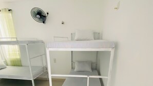 Ένα δωμάτιο με δύο κουκέτες, έναν ανεμιστήρα τοποθετημένο στον τοίχο και ένα παράθυρο με κουρτίνες.