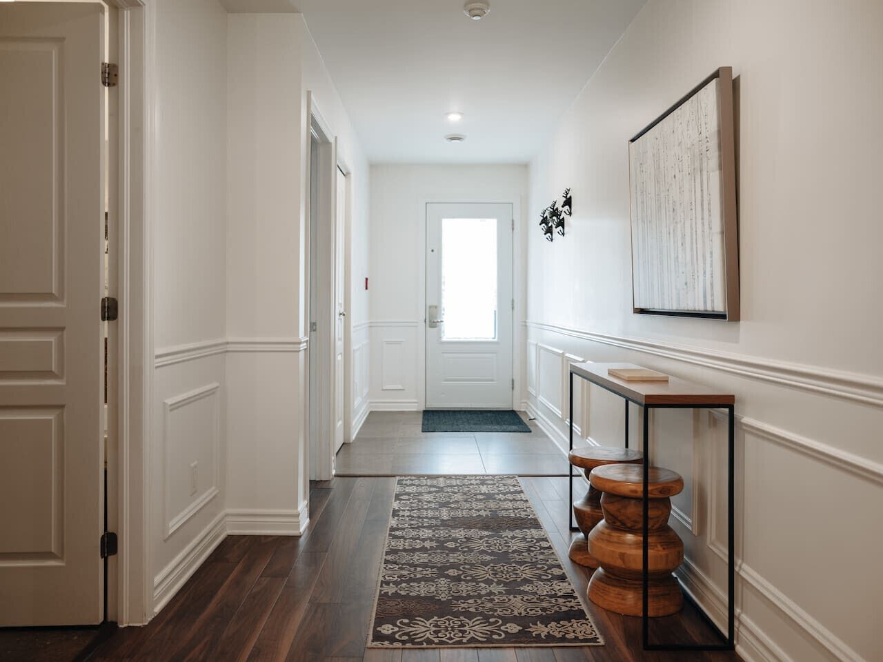 Un pasillo con piso de madera, un tapete estampado, una consola con taburetes de madera y una puerta que da a otra habitación.