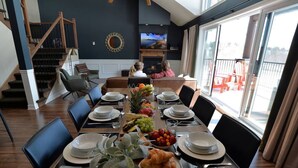 A dining area with a long table set for a meal, a TV mounted on the wall, and a view of the outdoors through large windows.