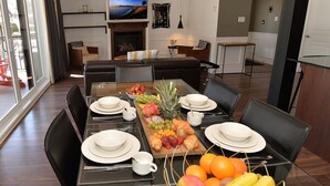 A dining area with a long table set for six, surrounded by black chairs. A television is mounted on the wall above the fireplace, and there are shelves with decorative items.