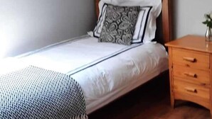 A single bed with a patterned pillow, a wooden nightstand with drawers, and a plain wall.