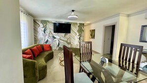A modern living room with a green sofa, a glass dining table, and a flat-screen TV mounted on the wall.