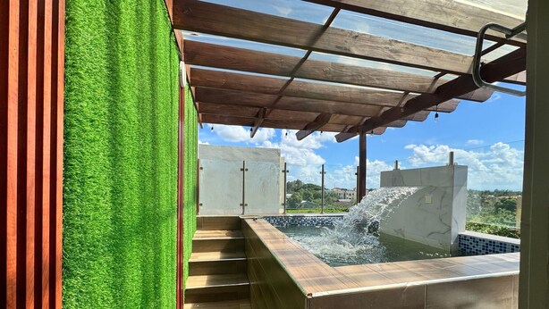 A modern outdoor hot tub area with a wooden pergola, artificial grass, and a view of a residential neighborhood.