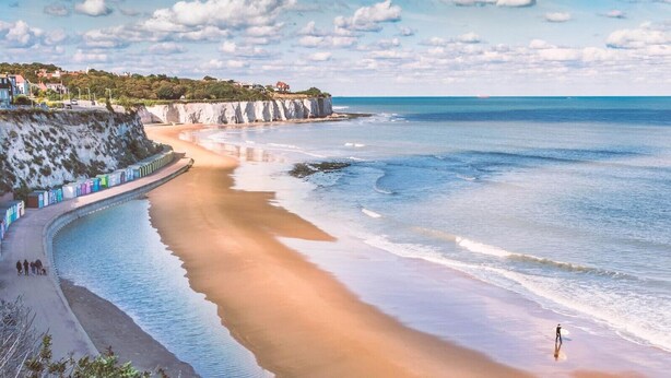 Una playa con una orilla curvada, una persona caminando y una fila de coloridas cabañas de playa a lo largo de la costa.