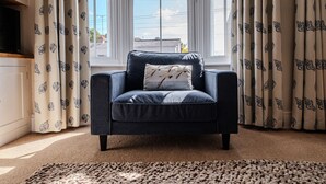 Un sillón azul oscuro con un cojín, frente a una ventana con cortinas.