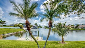 A serene lakeside view with two palm trees, a dock, and houses in the background. - Heated Pool - Dock - Gulf Access Canal - BBQ (Cape Coral)