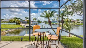 A covered outdoor area with a table and chairs, overlooking a canal with boats and palm trees. - Heated Pool - Dock - Gulf Access Canal - BBQ (Cape Coral)