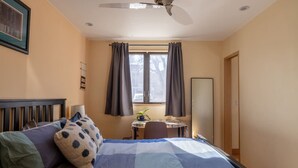 Une chambre à coucher comprenant un lit, un bureau, une chaise, un ventilateur de plafond et une fenêtre avec des rideaux.