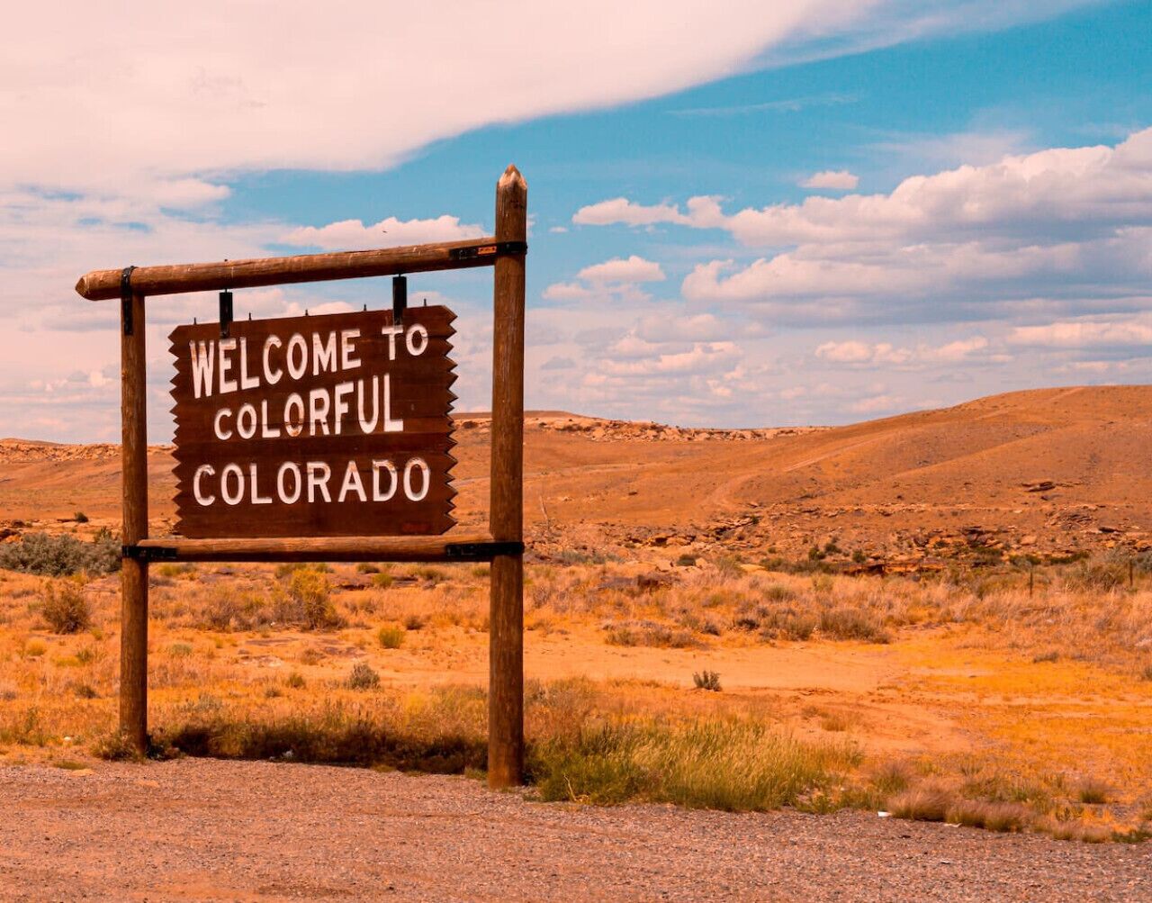 A wooden sign with the text 'WELCOME TO COLORADO' in a desert landscape.
