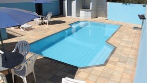 Una piscina rectangular amb una terrassa de rajoles, envoltada de cadires de plàstic blanc i un parasol blau.