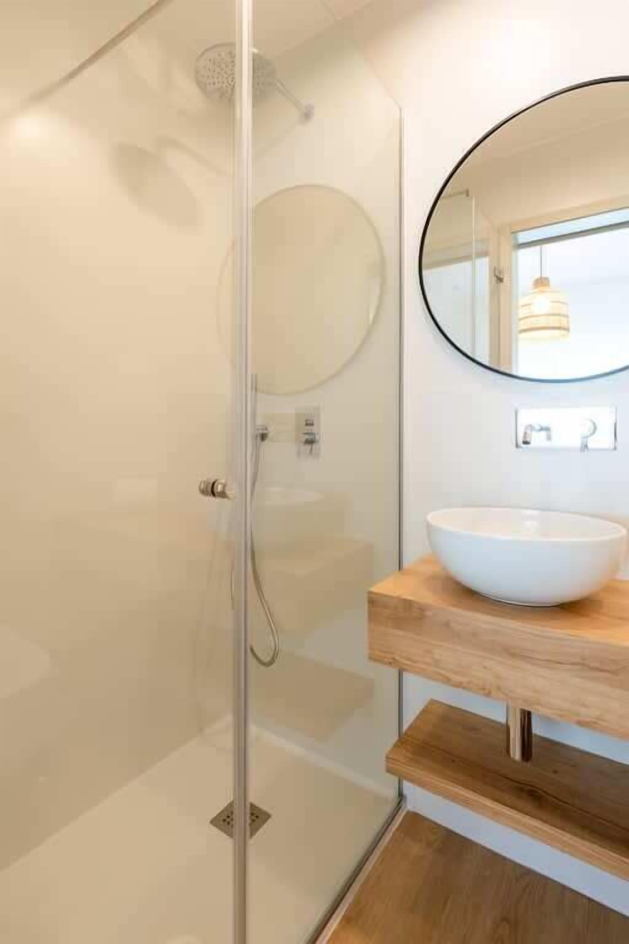 A modern bathroom with a glass-enclosed shower, a round mirror, and a wooden vanity with a white sink.