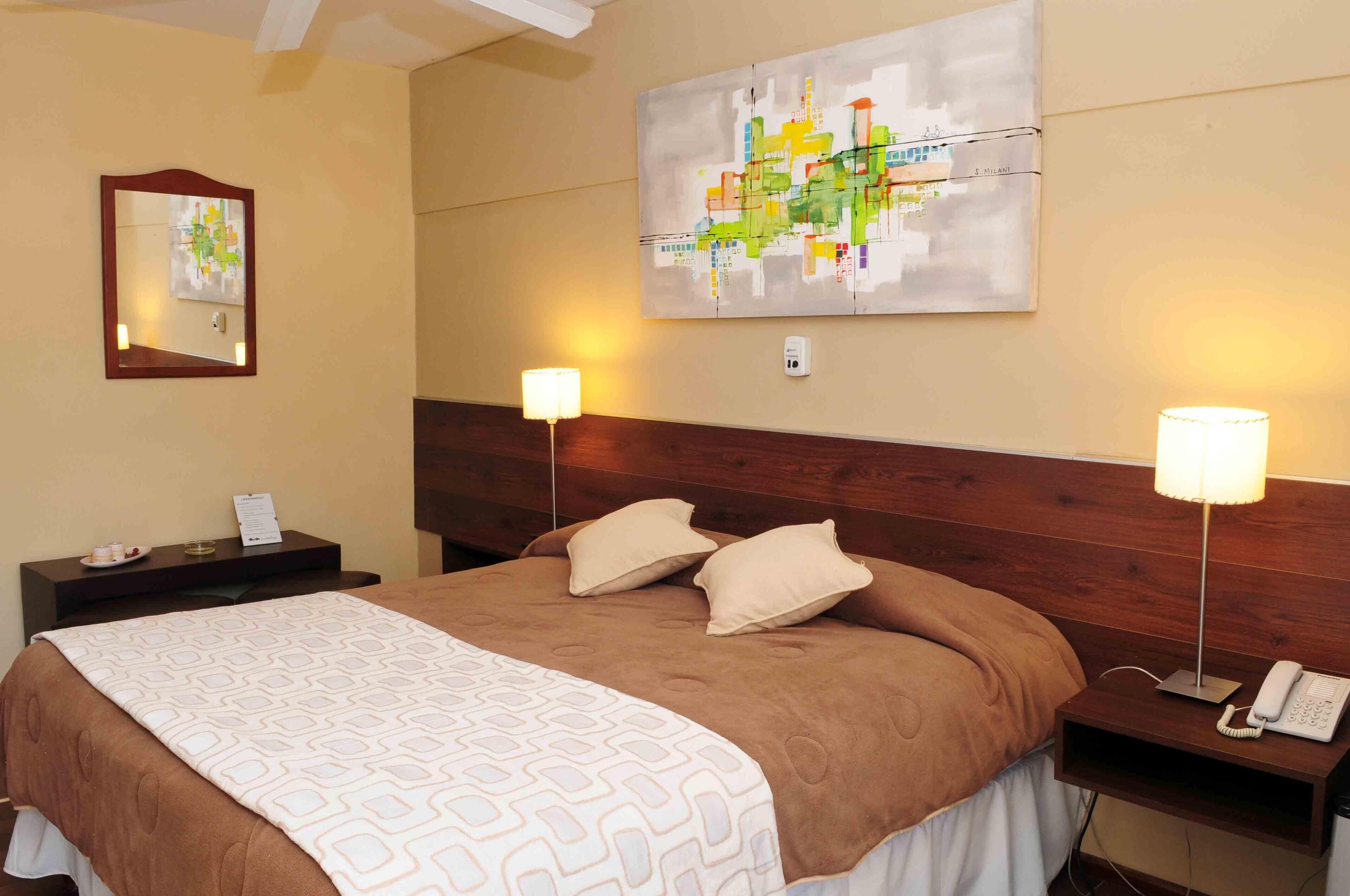 A hotel room with a large bed, two bedside tables, a nightstand, a lamp, and a painting on the wall.