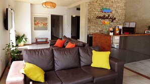 Un espace d’accueil avec un mur en pierre, un canapé en cuir marron agrémenté de coussins jaunes et orange, un banc en bois et un comptoir d’accueil sur lequel est apposée une enseigne indiquant « Catalujo ».