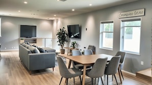 A modern living room with a grey sofa, a wooden dining table with chairs, a TV mounted on the wall, and a large window with a view. - 𝐵𝑒𝒶𝓊𝓉𝒾𝒻𝓊𝓁 Townhouse Haven from Hollywood—with PRIVATE BALCONY VIEW (Los Angeles)
