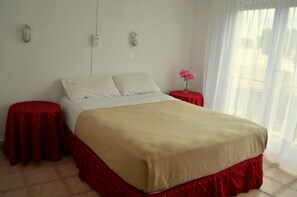 Ein ordentlich bezogenes Bett mit beiger Bettdecke und rotem Bettschlitten, zwei Nachttische in Rot, eine Vase mit rosa Blumen und ein Fenster mit transparenten Vorhängen.