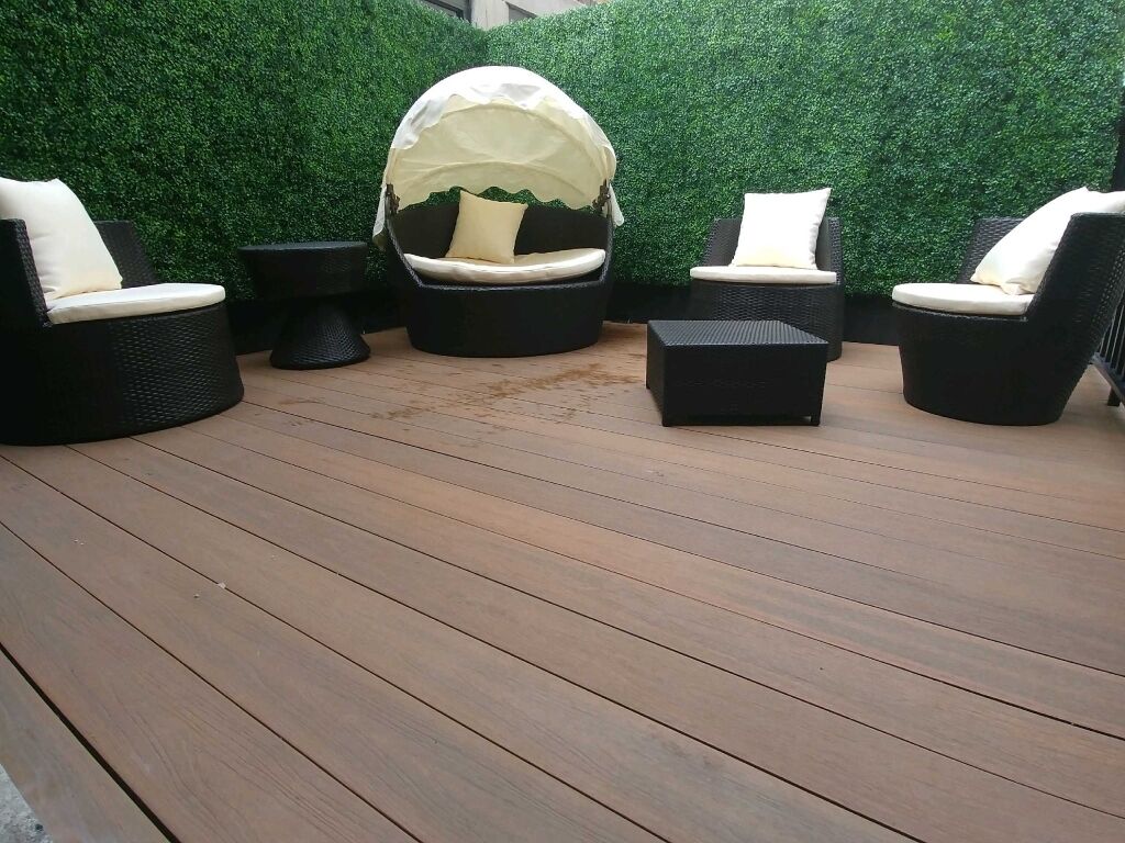 A patio with four black wicker chairs with white cushions, a round table, and a small square table. The floor is wooden, and the background features a green artificial hedge.