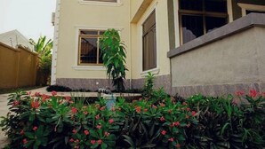 A two-story building with a beige facade, a small garden with red flowers, and a water feature. - Salma's Place (Kumasi)