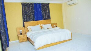 A bedroom with a wooden bed, white bedding, a nightstand with a lamp, and a patterned curtain. - Salma's Place (Kumasi)
