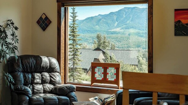 A room with a leather armchair, a wooden table, and a view of mountains through a large window. - The Barn: Private Deck & Mountain Panoramas (Fernie)