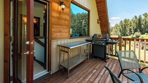 A wooden deck with a grill and a table, a door leading inside, and a view of trees. - The Barn: Private Deck & Mountain Panoramas (Fernie)