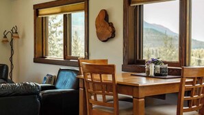 A dining area with a wooden table and chairs, a wooden wall decor, and a view of mountains through large windows. - The Barn: Private Deck & Mountain Panoramas (Fernie)