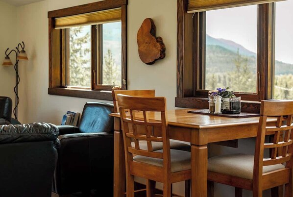 A dining area with a wooden table and chairs, a wooden wall decor, and a view of mountains through large windows. - The Barn: Private Deck & Mountain Panoramas (Fernie)
