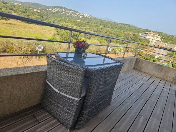 Varanda com móveis de vime e mesa de vidro, com vista para uma paisagem verde e edifícios.