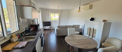 A modern kitchen with dark wood cabinets, a built-in stove, and a sink. There is a dining area with a round table and two chairs. A sofa and a TV are also visible.
