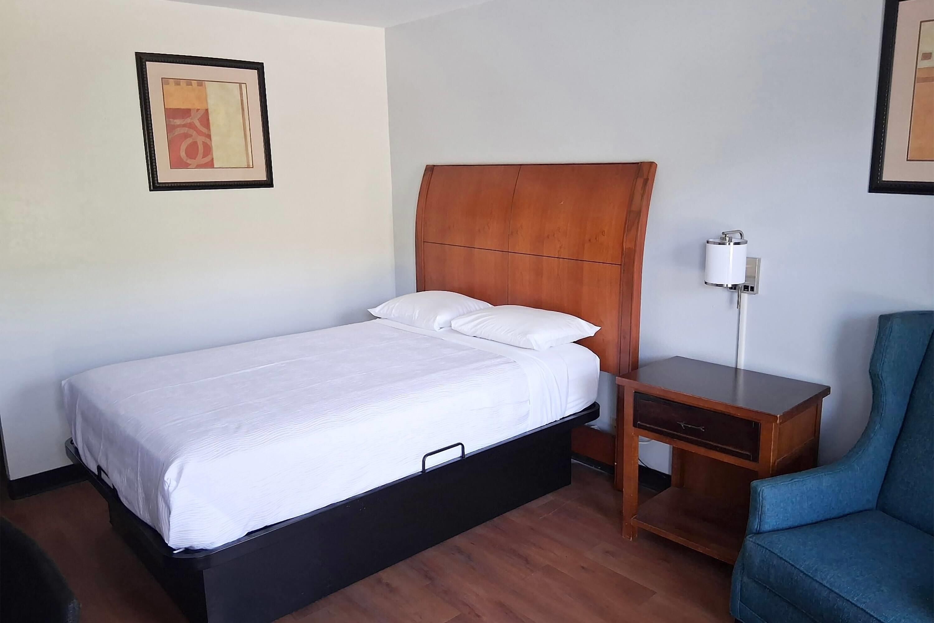 A hotel room with a single bed, a wooden headboard, a bedside table with a lamp, and a blue chair.
