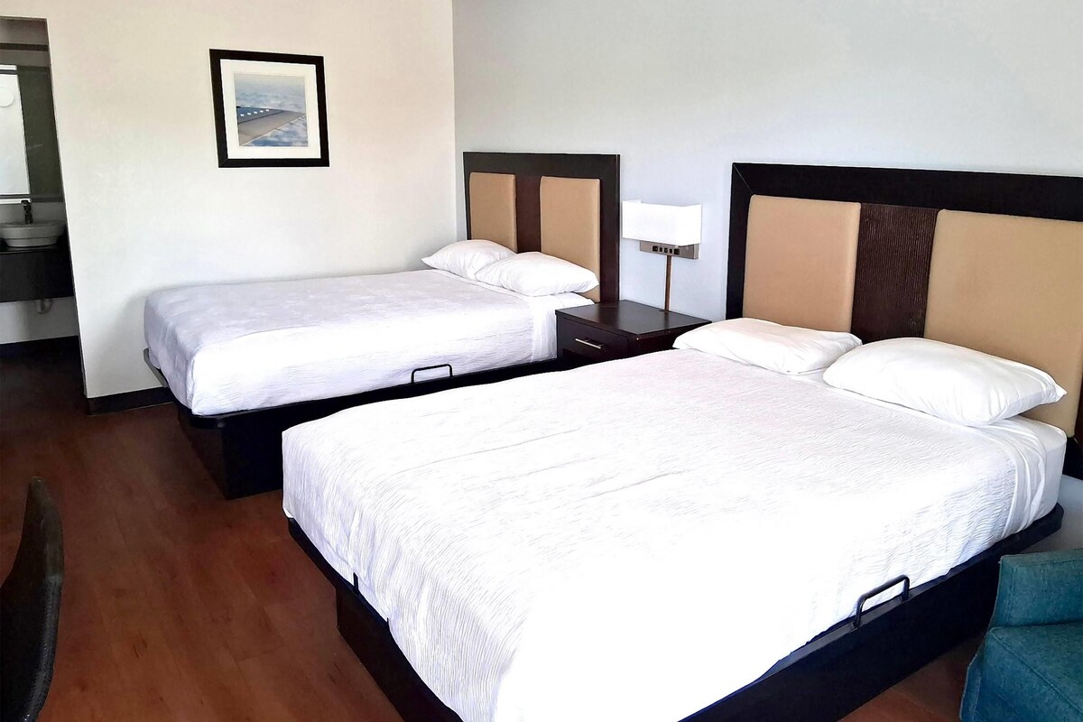 A hotel room with two beds, a bedside table with a lamp, a framed picture on the wall, and a wooden floor.