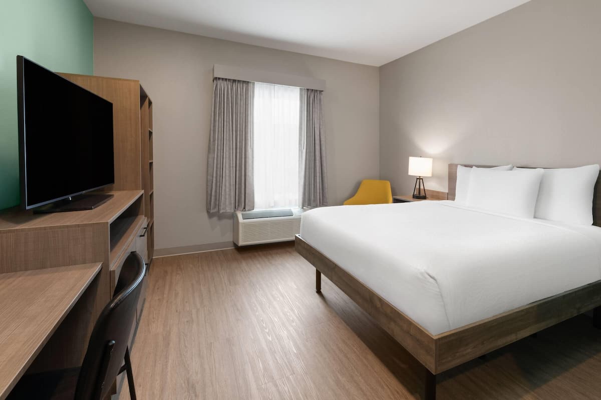 A hotel room with a bed, a desk with a TV, a chair, a lamp, and a window with curtains.