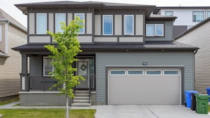 A two-story residential house with a garage, a small porch, and a front yard with a young tree. - Cozy 2BR/2BA Suite | 10 Mins to Airport | Private Entry & Kitchen (Calgary)