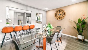 Uma sala de jantar moderna com mesa de vidro, cadeiras laranja e relógio de madeira na parede.