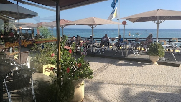 Un espace extérieur aménagé avec des tables et des chaises sous des parasols, offrant une vue sur une plage où sont amarrés des bateaux.