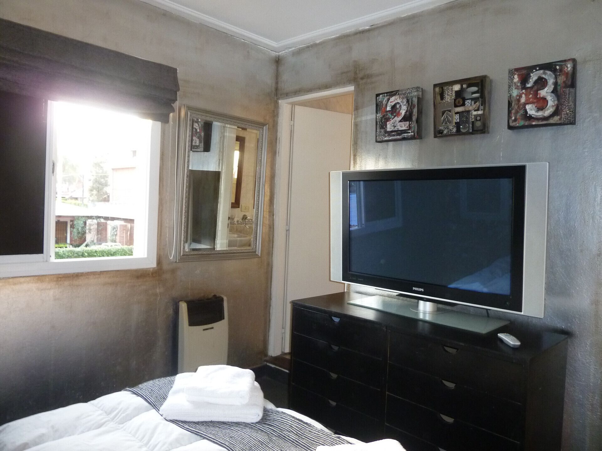 Un dormitorio con una televisión de pantalla plana grande, una cama con ropa blanca, un aparador y una ventana que ofrece vistas a un edificio.