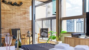 Una camera d'albergo moderna con un'ampia finestra, un letto con asciugamani ordinati, un tavolino con una bottiglia e dei bicchieri, e una libreria piena di libri.