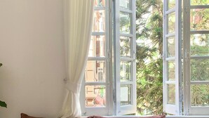 A brown sofa with a white pillow, a round wooden table with a magazine, and a window with open shutters. - 2 Bedrooms in a Cozy and Aesthetic Villa / Naturally Cool / West Lake / Kotton 2 (Hà Nội)