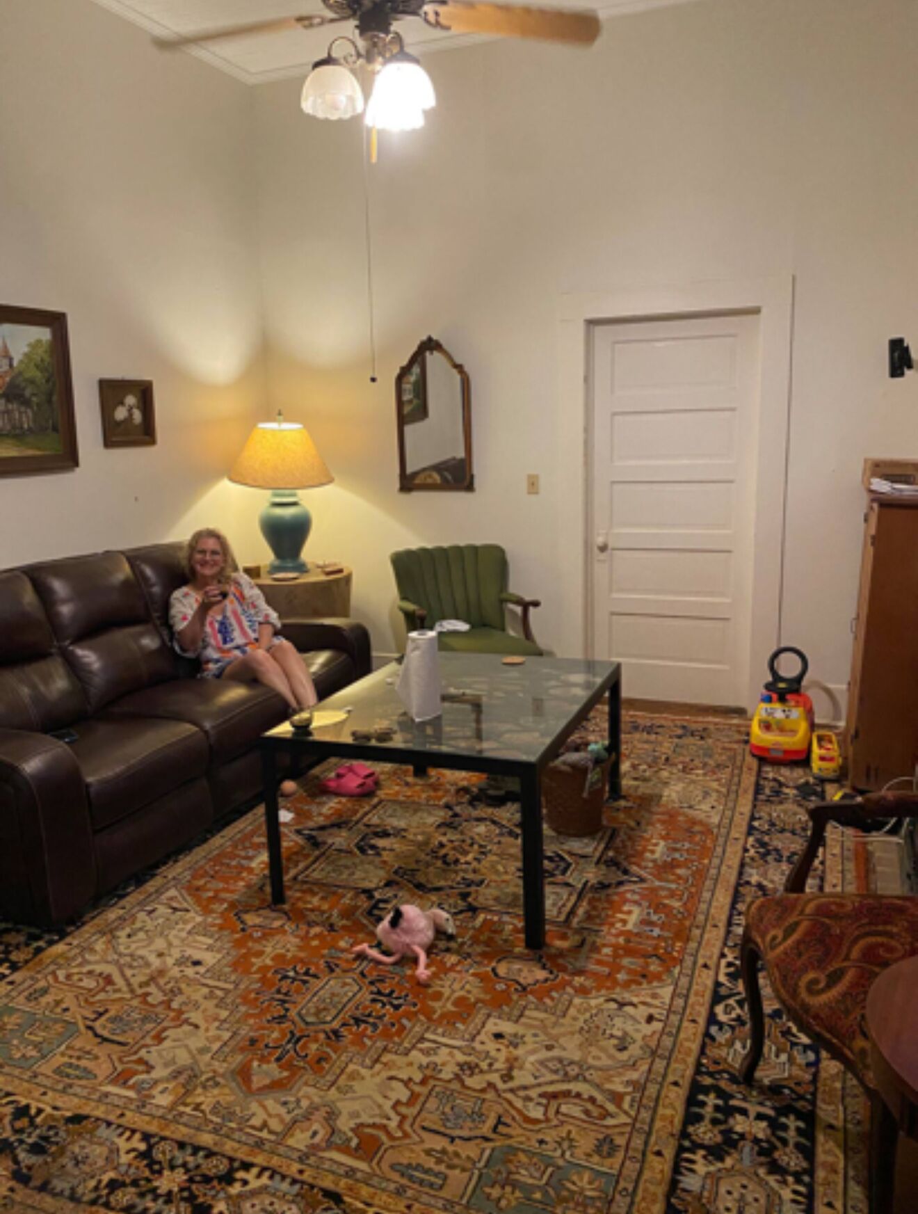 A living room with a brown leather couch, a glass coffee table, a patterned rug, a green armchair, and a ceiling fan.